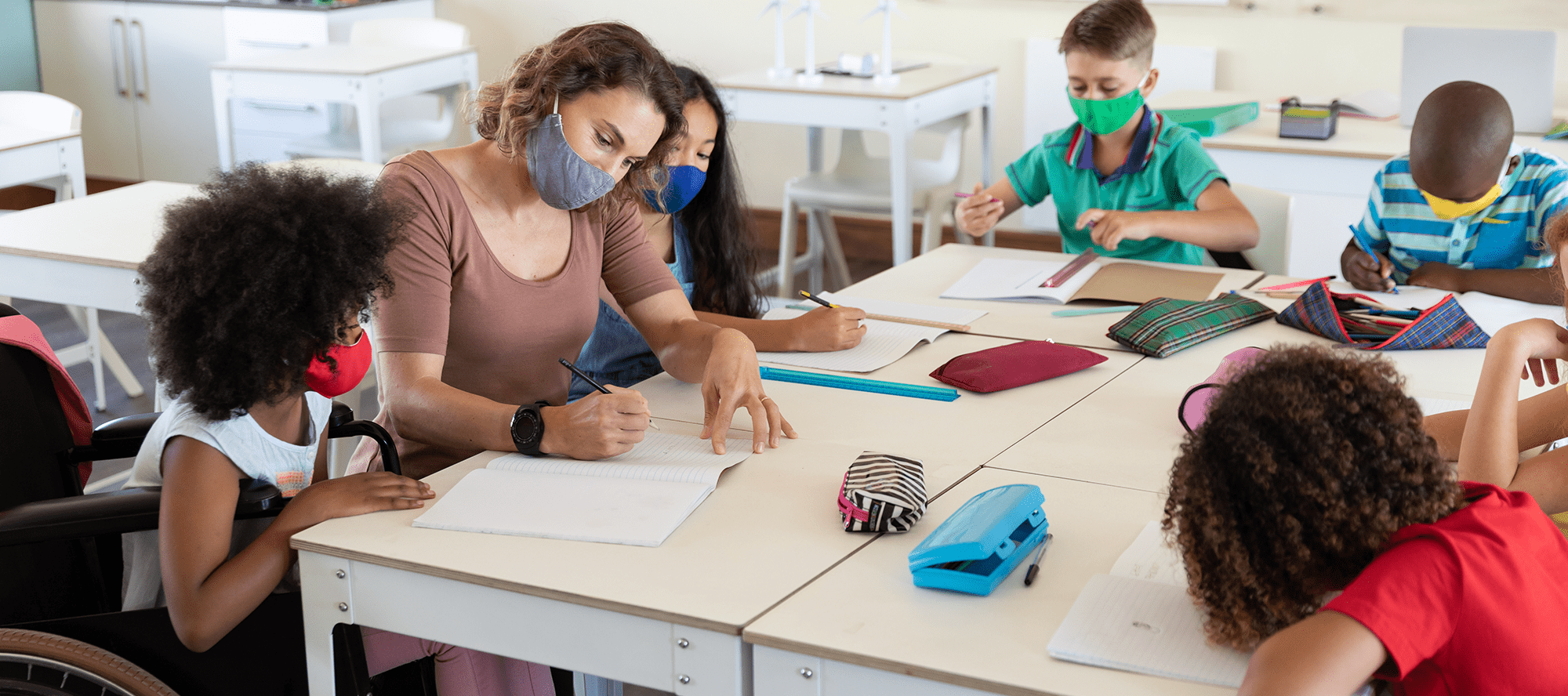 Teacher sitting at a table with students, all wearing masks.