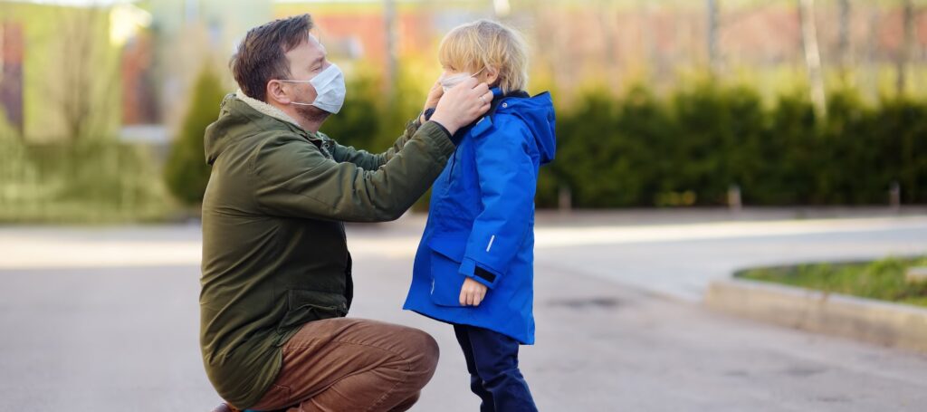 Mature man wearing a protective mask puts a face mask on a his son in airport