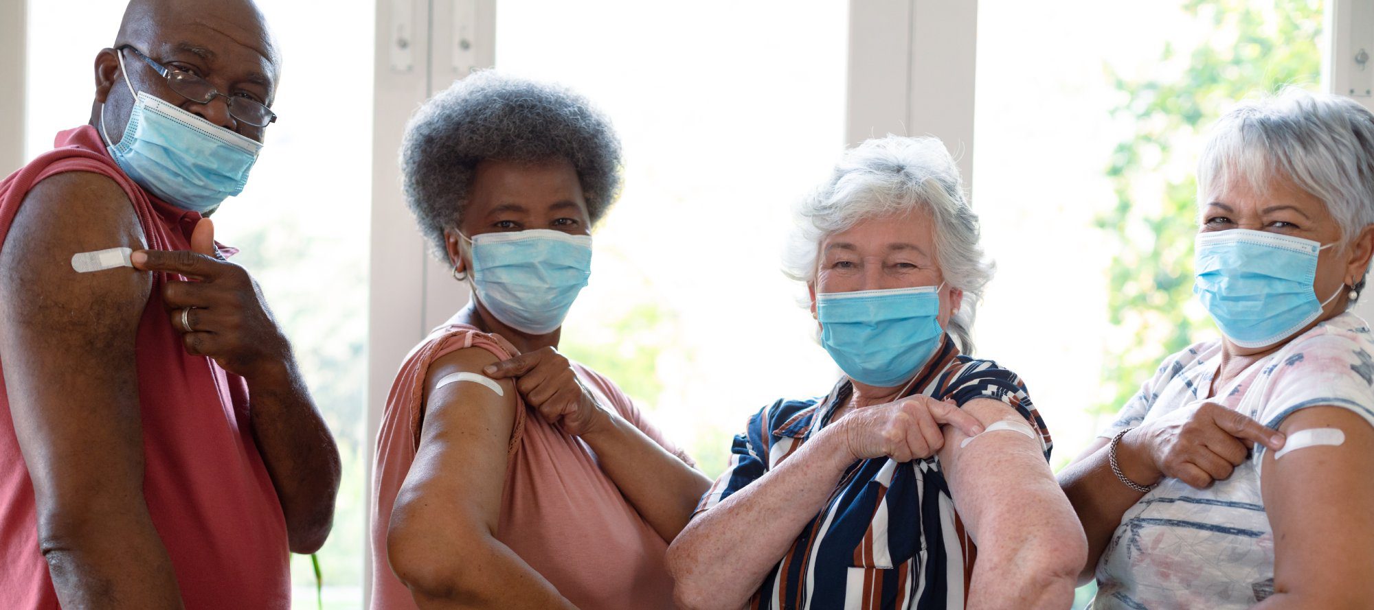 Four diverse male and female senior friends wearing face masks showing plasters after vaccination