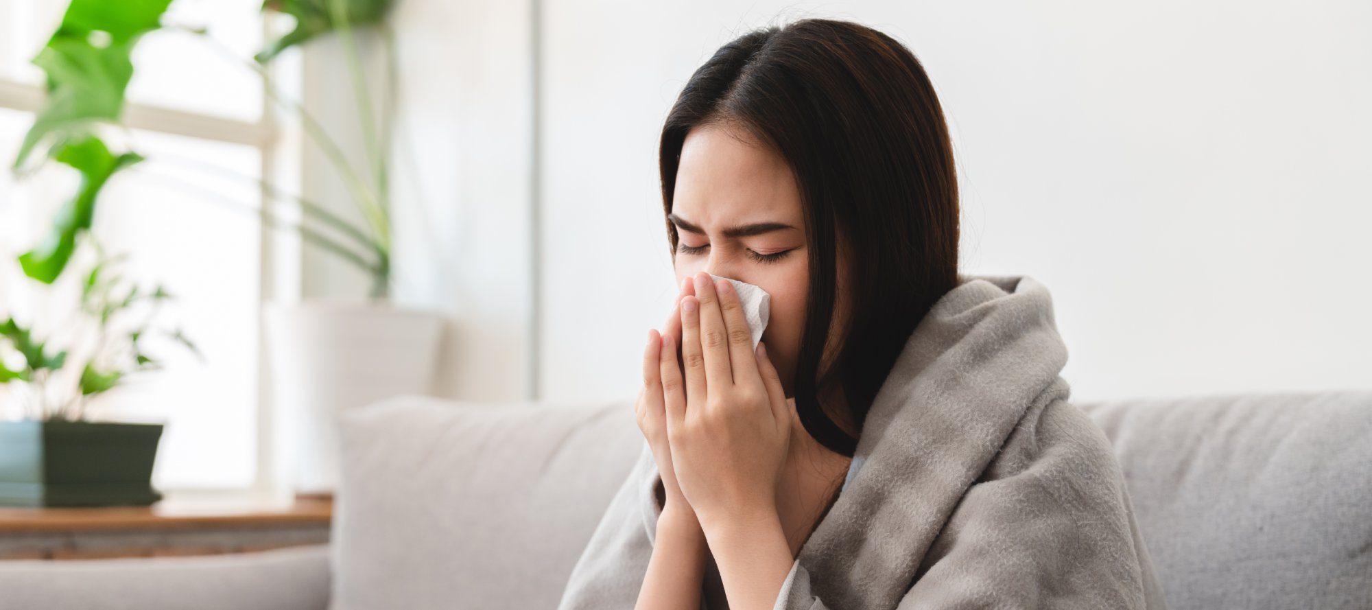 sick asian young woman sneezing into tissue paper while covered with a blanket