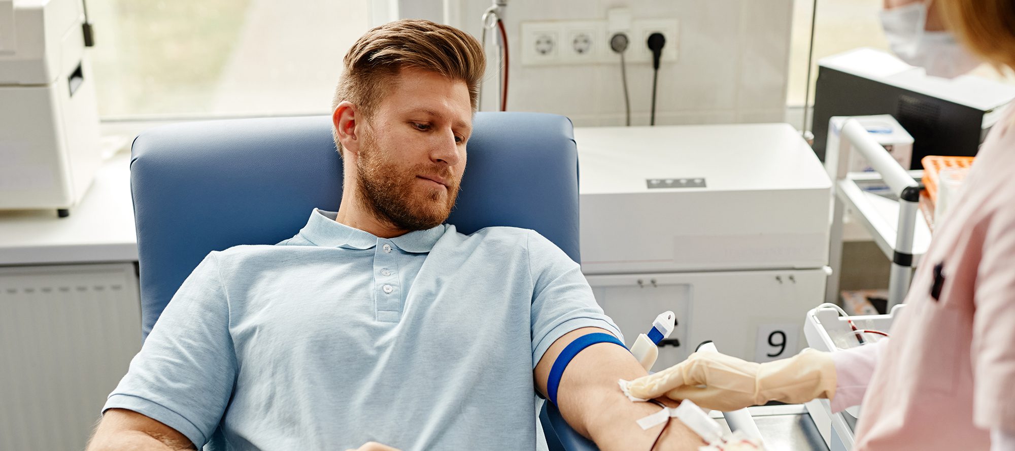 Portrait of handsome young man preparing for blood donation at med center with nurse helping