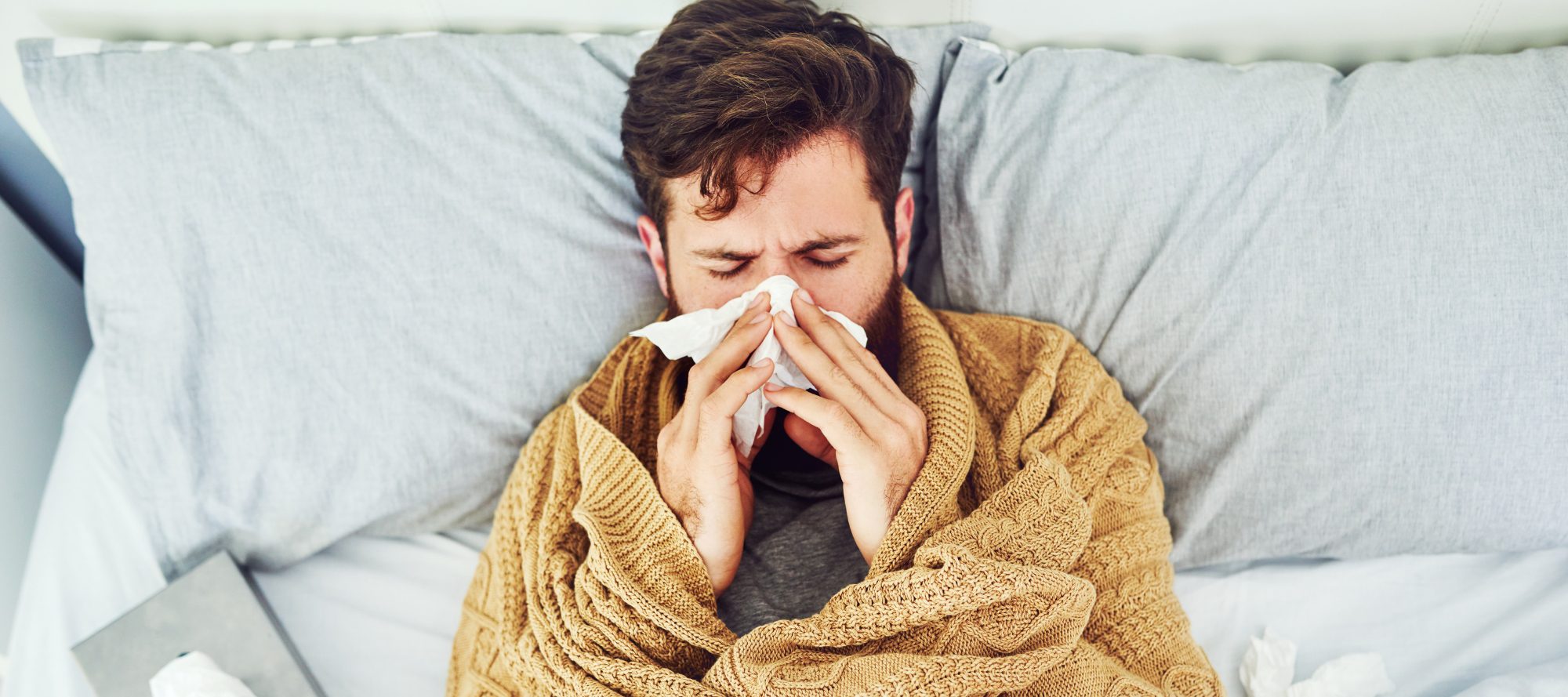 Man laying in bed wrapped in blanket, resting his head on pillow as he blows his nose into a tissue
