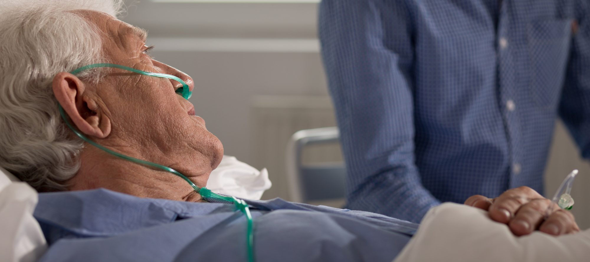 A hospitalized elderly man laying in bed with additional oxygen being provided to him while someone sits next to the bed